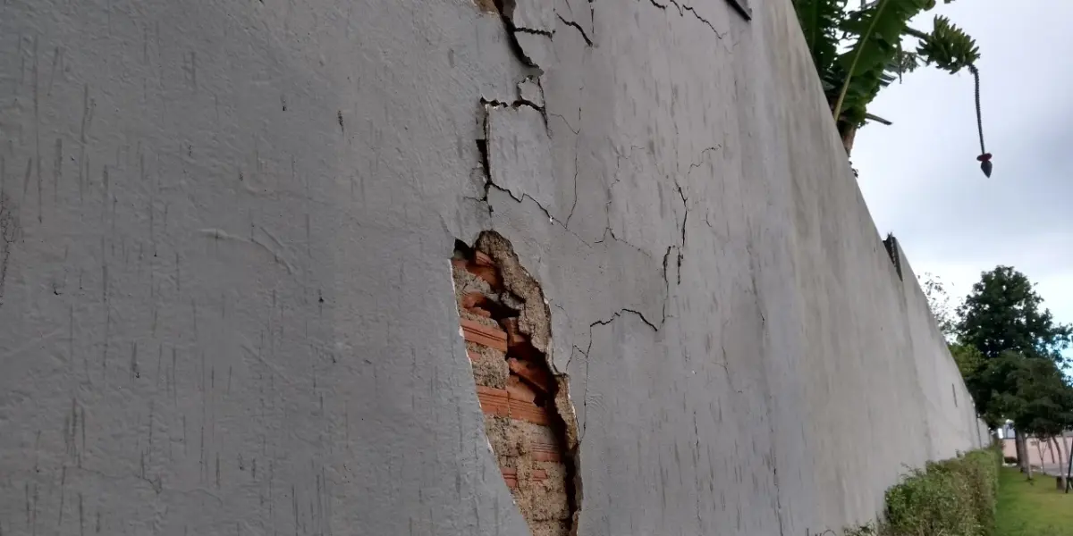 Muro de alvenaria com o reboco destacado e tijolos expostos, ao lado de fissuras no revestimento