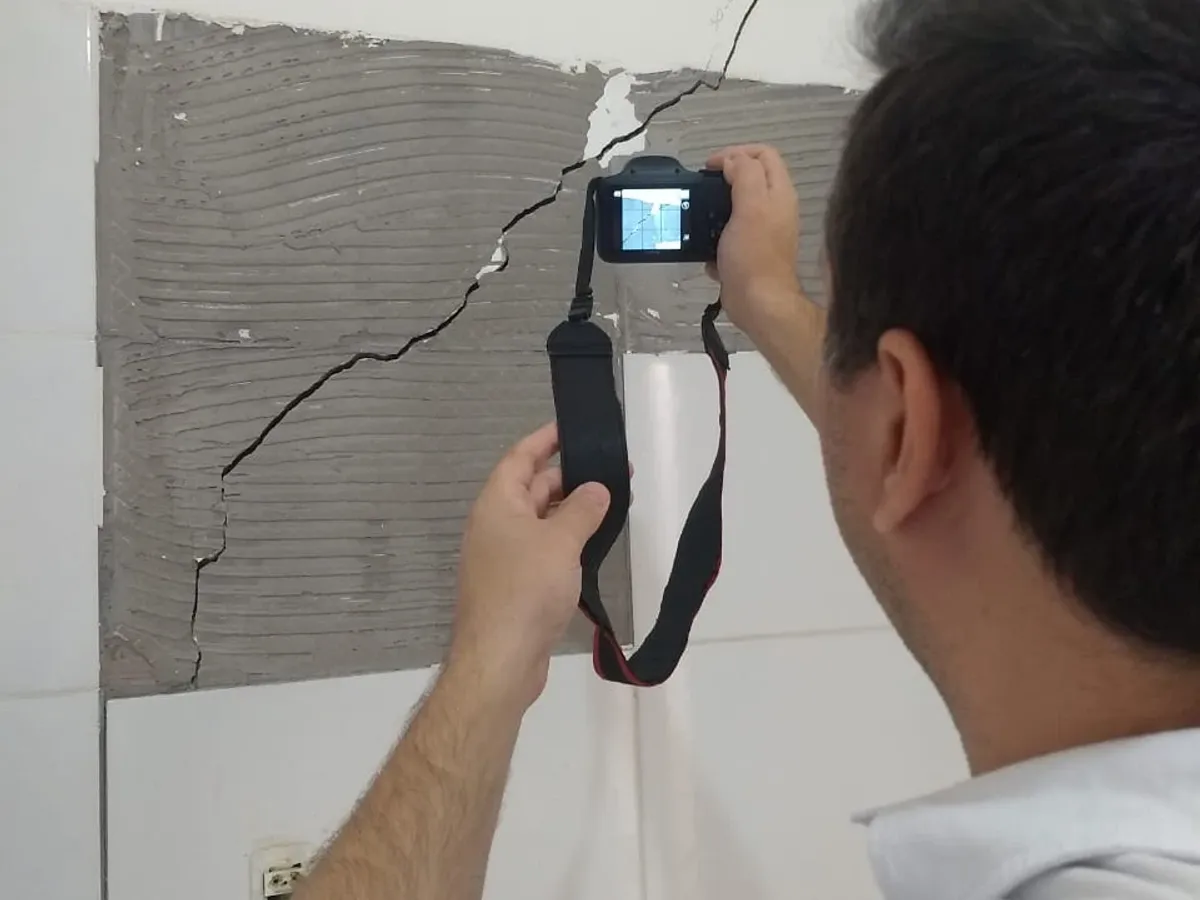 Vistoriador de camisa polo branca, de costas, fotografa com câmera digital a trinca que atravessa a parede no trecho onde o revestimento cerâmico se soltou