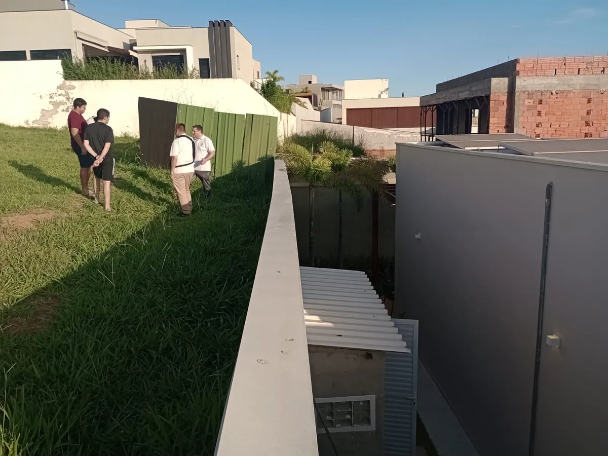 Engenheiros conversam com moradores num terreno em aclive, junto ao muro de divisa, com a obra do lote vizinho ao fundo